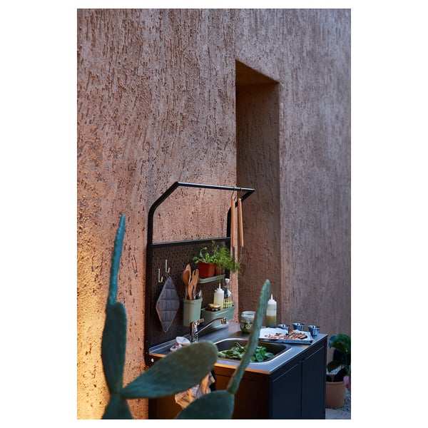 Outdoor kitchen setup with stainless steel sink and galvanized steel shelves against textured wall. Various kitchen tools and plants arranged neatly.