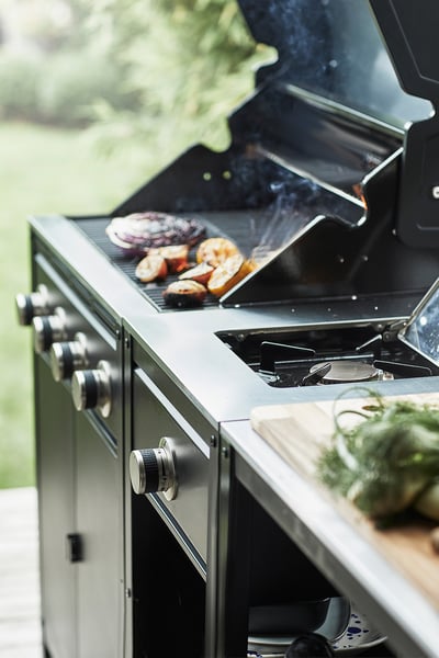 Stainless steel outdoor grill with vegetables cooking, side burner, green lawn background.