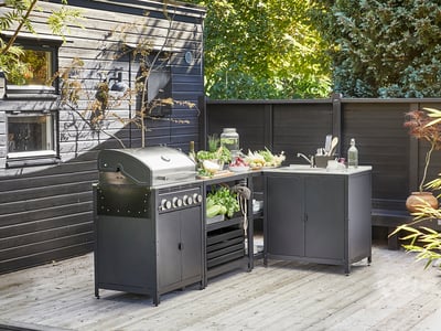 Outdoor kitchen setup with dark gray grill, counter, and sink on deck, surrounded by greenery.