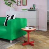 Green sofa, black/white pillow, red side table with mug, glasses, books. Storage with plant. Pink walls, art. Wood floor, grey rug.