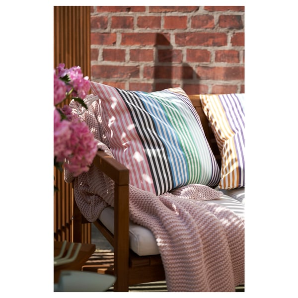 Colorful striped outdoor cushions on wooden bench with pink flowers and brick wall.