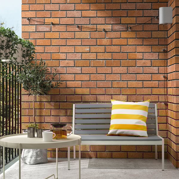Cozy balcony with grey bench, yellow cushion, round table, teapot, cups, plant, olive tree, brick wall.