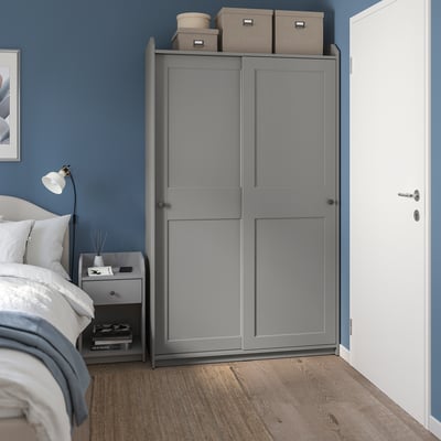 Minimalist blue bedroom with gray HAUGA wardrobe, wooden floor, white bed, and nightstand with lamp.