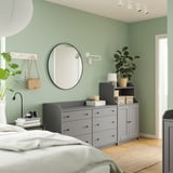 Bedroom with light green walls, wide gray HAUGA dresser, round mirror, plants, white bed.