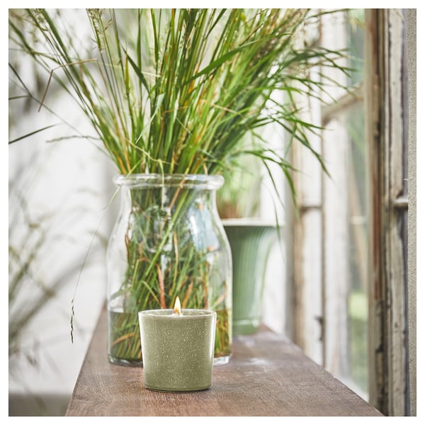 Green scented candle on wooden shelf near glass vase with greenery.