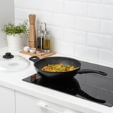 Black wok with veggies cooking on modern stove, sturdy handles, glass lid, nearby herbs.