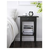 Black HEMNES nightstand with books, cup, and alarm clock beside large bed.