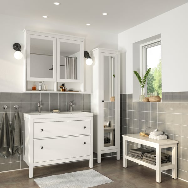 Modern white bathroom with HEMNES vanity, wide mirror cabinet, tall storage unit, and bench. Dark grey tiles, oval basins, and round lights.