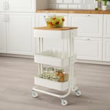 White kitchen cart with wheels, holding a glass bowl of salad on a bamboo chopping board, stacks of cups, and jars with snacks.