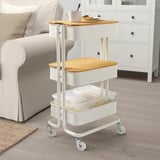 Small kitchen cart with wooden top, clear container, and beige cushioned chair.