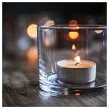 Small candle in clear glass holder; lit wick, soft glow, reflective surface.