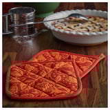 Two red-orange patterned potholders on wooden table, with flour sifter and pie dish nearby.