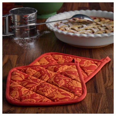 Two red-orange patterned potholders on wooden table, with flour sifter and pie dish nearby.
