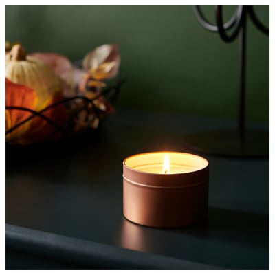 A lit candle in a tin on a black surface, with autumn decorations in the background. It releases a calming scent of sandalwood, patchouli, and fir.
