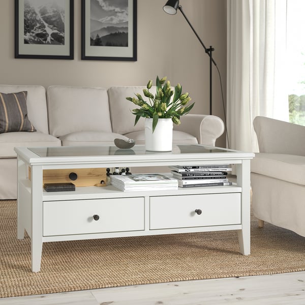 White IDANÄS coffee table with glass top, drawers, and flowers. Centered in a living room with a beige sofa and rug.
