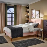 Bedroom with IDANÄS pink bed, dark quilt, white pillows, curtains, wooden furniture, floor rugs, and window view.