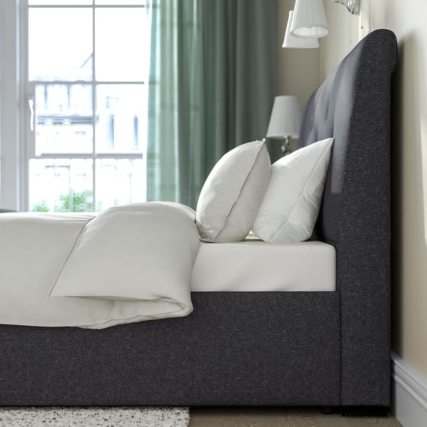 Cozy bed with sloped headboard, soft upholstery, and under-bed storage, featuring white pillows and lamps beside a window with curtains.
