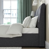 Cozy bed with sloped headboard, soft upholstery, and under-bed storage, featuring white pillows and lamps beside a window with curtains.