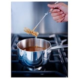 A hand uses a balloon whisk to stir liquid in a saucepan on a gas stove. The whisk is held by a long handle, ensuring safe mixing over the heat.