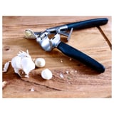 Garlic press with black handle on wooden table, with garlic cloves and peels.