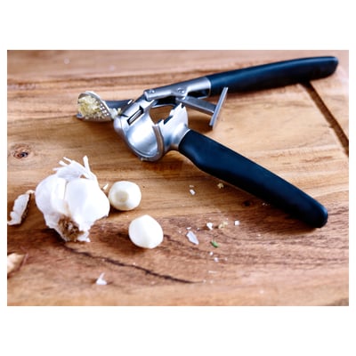 Garlic press with black handle on wooden table, with garlic cloves and peels.