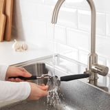 Person presses garlic under running water from a stainless steel faucet in a kitchen sink.