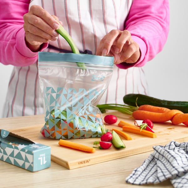Person sealing a reusable vegetable bag with colorful pattern on a wooden table surrounded by fresh produce.