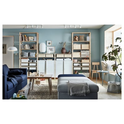 Modern living room with blue couch, gray ottoman, wooden shelves full of books and decorative items, large window, and blue walls.