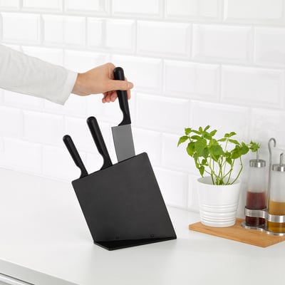 Person grabbing knife from black block on kitchen counter, beside plant and condiments.
