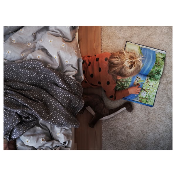 Child in orange polka dot shirt lies near bed reading colorful book with dinosaurs, beside a stuffed dinosaur toy, on a grey rug.