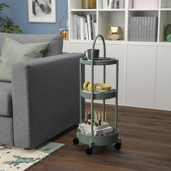 Small green 3-tier rolling trolley beside large grey couch, holding mugs and books.