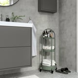 A grey-green, three-tier rolling cart named JUTTERSBO next to a bathroom sink. It holds towels and toiletries, fitting well in small spaces.