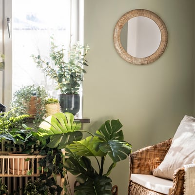 Round rattan mirror with japandi style above wicker chair, green wall, plants.