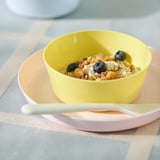 Bright yellow bowl holds creamy oatmeal topped with fresh blueberries, placed on a pink plate with a spoon.