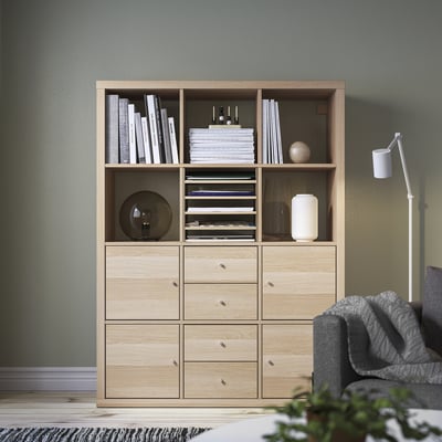 KALLAX light wood shelf unit with books, decor, and drawers against wall, gray sofa right.