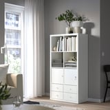 A white KALLAX shelf unit in a bright room with an open shelf displaying books and decorative items, and a cabinet with a lock for secure storage.