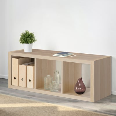 Wooden KALLAX shelving unit with storage boxes, vases, and plant on top, against a wall. Used for display and storage.