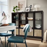 Modern dining area with black KALLAX shelving unit, displaying plates, vases, and a lamp. Two blue chairs and a woman are visible.