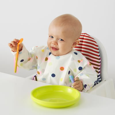 Baby sits in highchair wearing colorful bib with front pocket holding orange spoon and blue baby utensil over green plate.