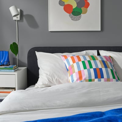 Cozy bedroom with black bed, pastel checkered pillow, abstract art, and nightstand with lamp and plant.