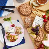 Top view of a snack setup with KNÄCKEBRÖD RÅG crispbread, cheeses, crackers, fruit, and radishes on a wooden board and plate.