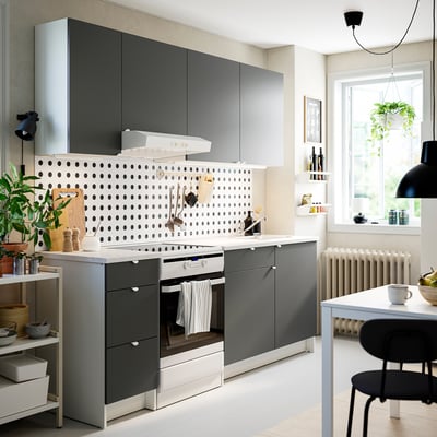 Modern kitchen with grey cabinets, white counters, and polka-dot backsplash. Black chairs by window, plants, and decor add warmth.
