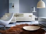 Modern living room with white sofa, white chair, hide rug, and woven pendant lamp. Blue walls and minimalist design.