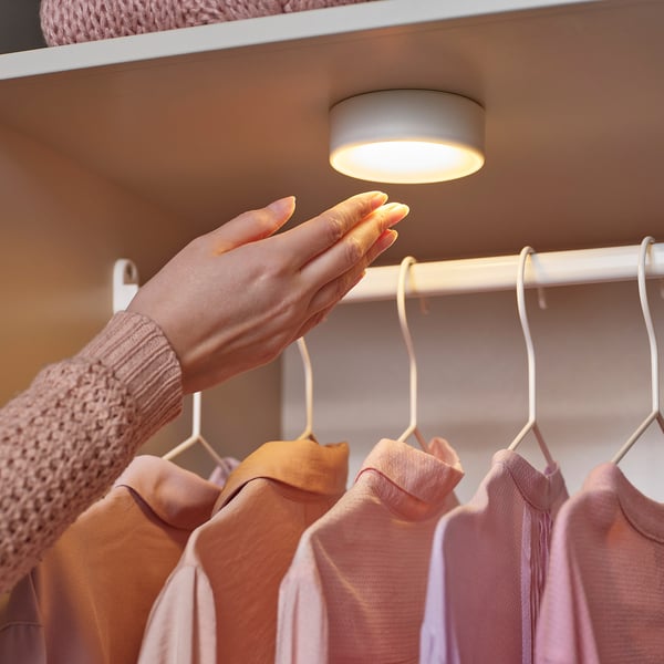 A hand pressing a cordless closet light, illuminating shirts on hangers beneath a shelf.