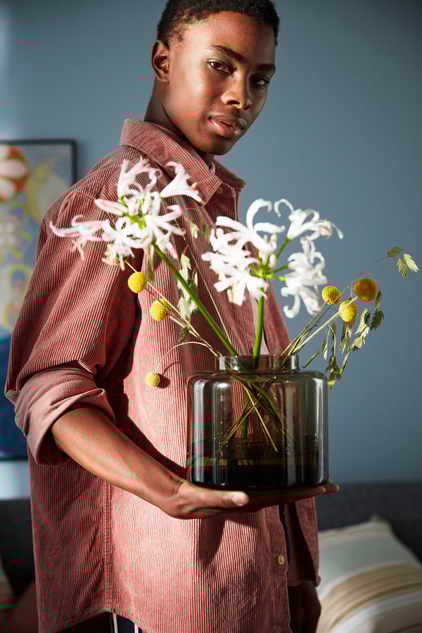 A person holds a transparent brown glass vase with white flowers and small yellow spheres, part of the KONSTFULL series designed by ilse crawford.
