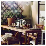 Outdoor table set with wooden chairs, KORKEN jars, glasses, plates, cutlery, bowls of fruit, and string lights.