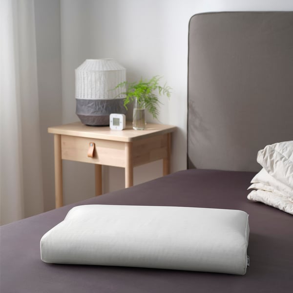 Modern bedroom with gray headboard, wooden nightstand, lamp, clock, green plant, and ergonomic pillow.