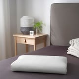 Modern bedroom with gray headboard, wooden nightstand, lamp, clock, green plant, and ergonomic pillow.