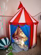 Bright kids play tent shaped like a circus tent, red and white striped top with blue walls, cozy inside with pillows and a small lamp.