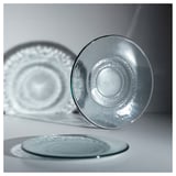 Three transparent glass plates in various positions on a gray surface, showcasing their unique, textured design and recycled glass composition.